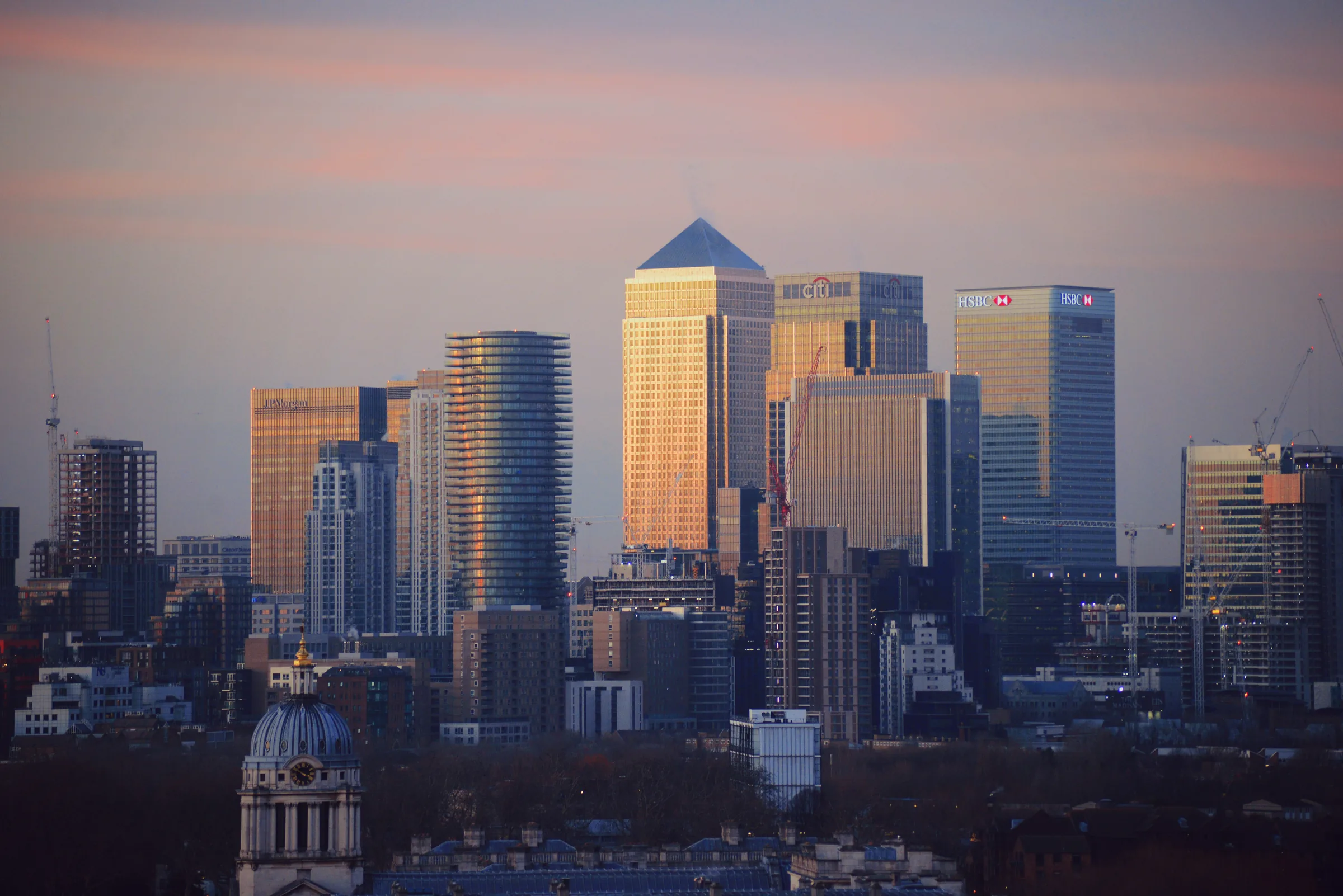 Canary Wharf skyline at sunset showing One Canada Square, HSBC, Citi and JPMorgan towers, with the Old Royal Naval College dome in the foreground